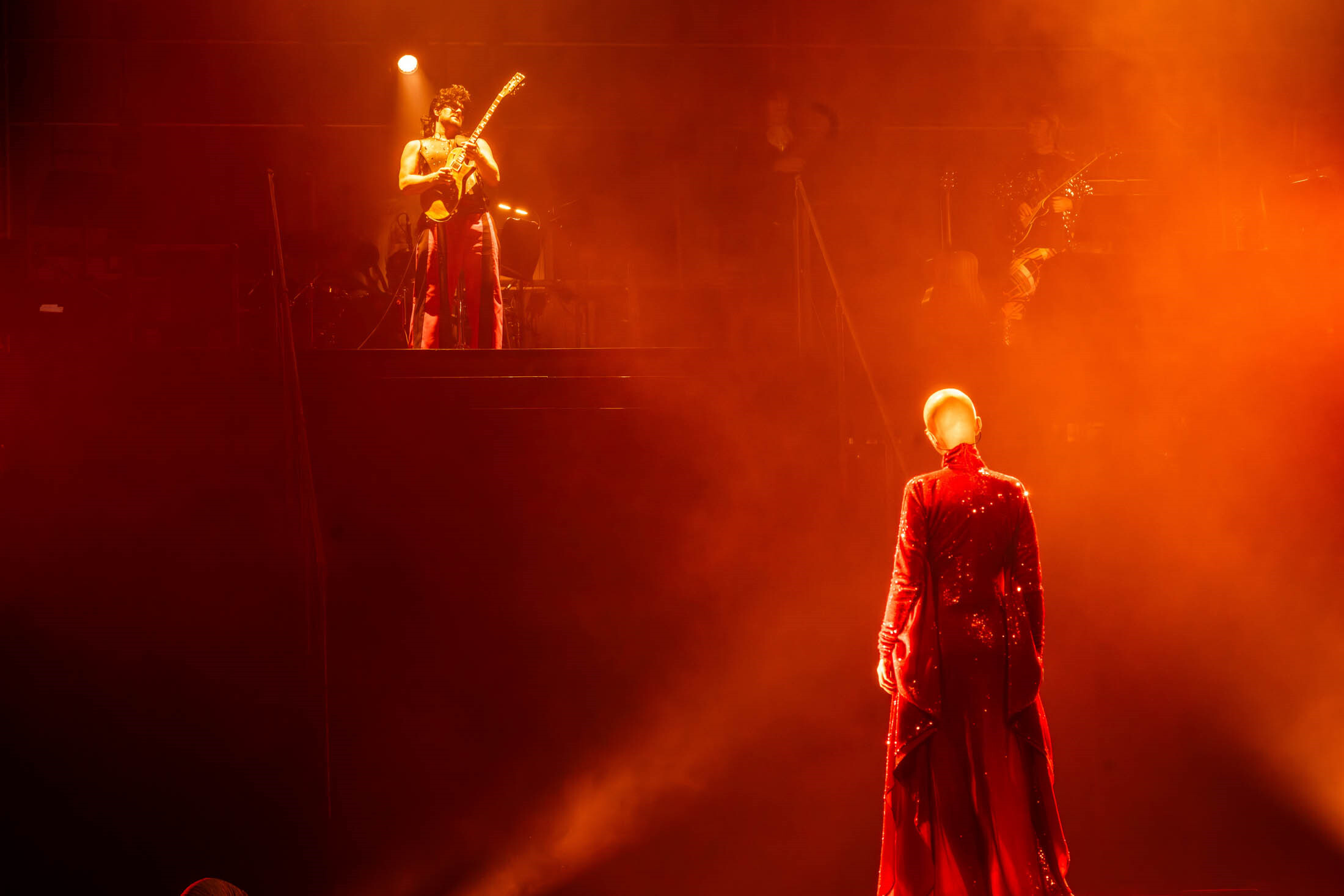 Bühne in rotes Licht getaucht, Sängerin in rotem Pailettenkleid blickt auf einen Rock-Gitarristen auf einem hohen Podest über ihr