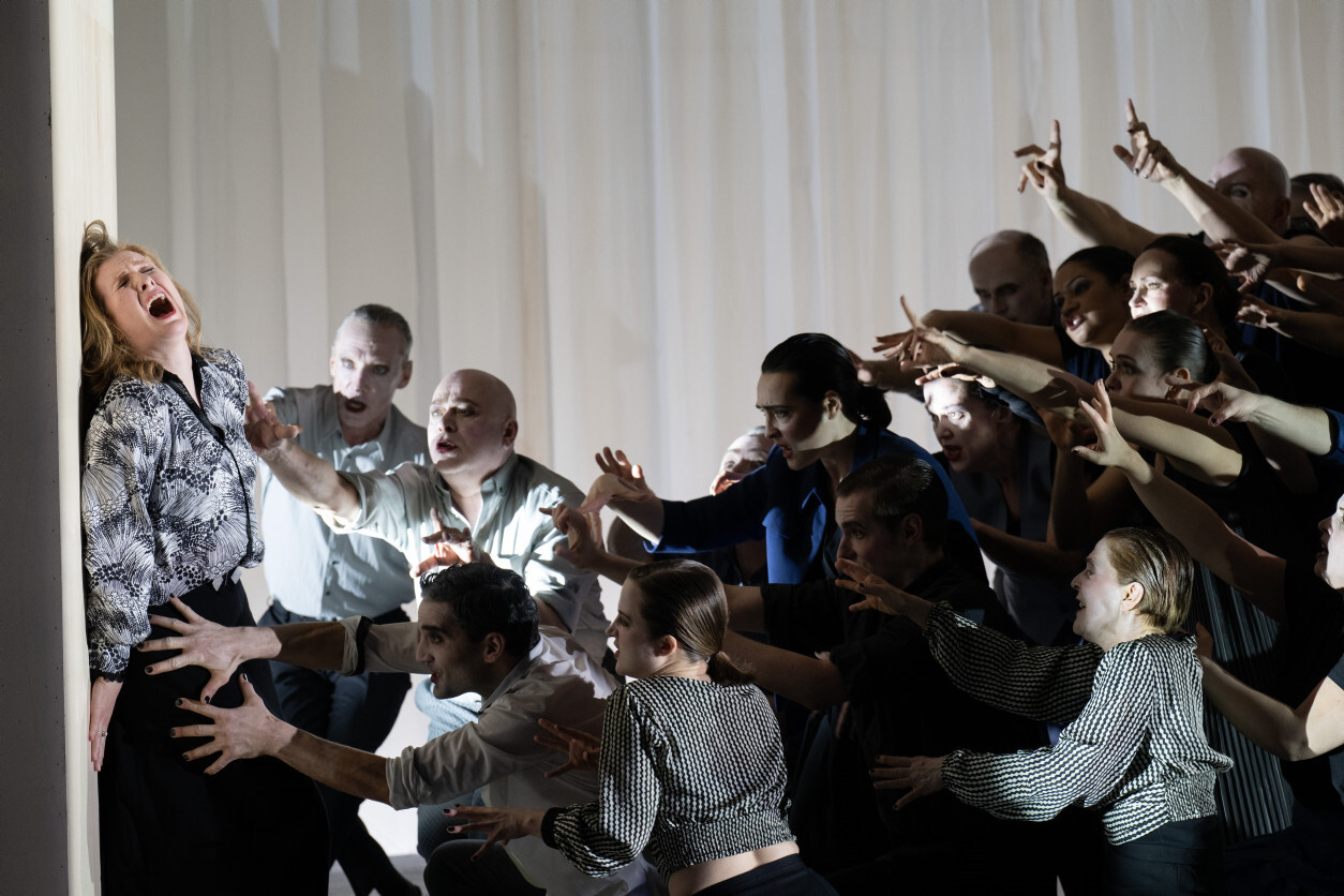 Paula Murrihy als Dejanira links an Wand stehend bedrängt von einer Gruppe Tänzer:innen und Sänger:innen