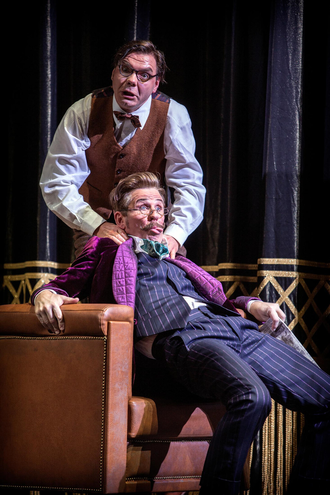 Zwei Darsteller sitzen gemeinsam auf einem Sessel, der eine mit einer Zeitung in der Hand