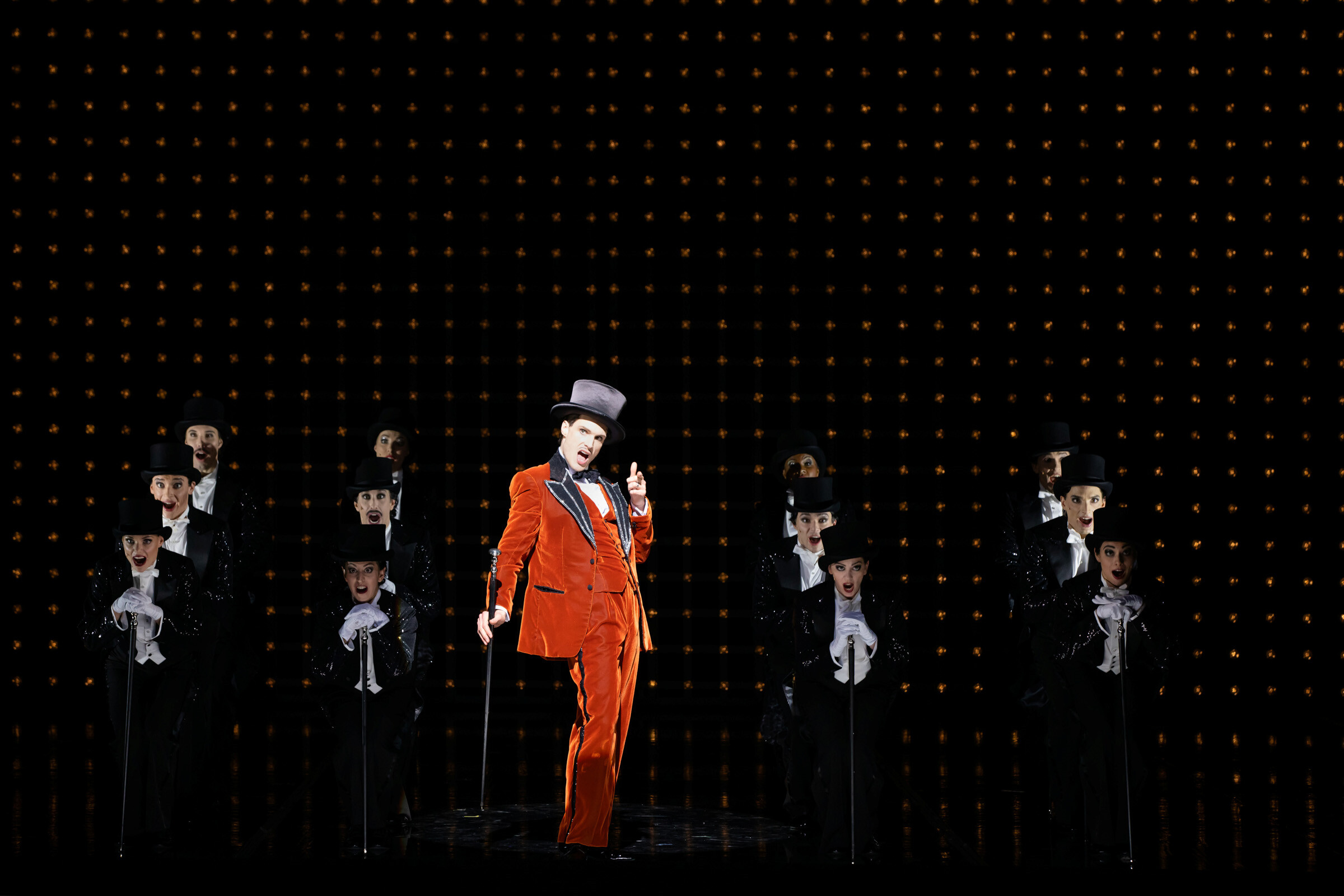 Jörn-Felix Alt als Billy Flinn in orangenem Anzug und Zylinder sowie Tänzer:innen in schwarzen Anzügen und mit Zylinder auf dem Kopf auf  Bühne Komische Oper Berlin im Schillertheater